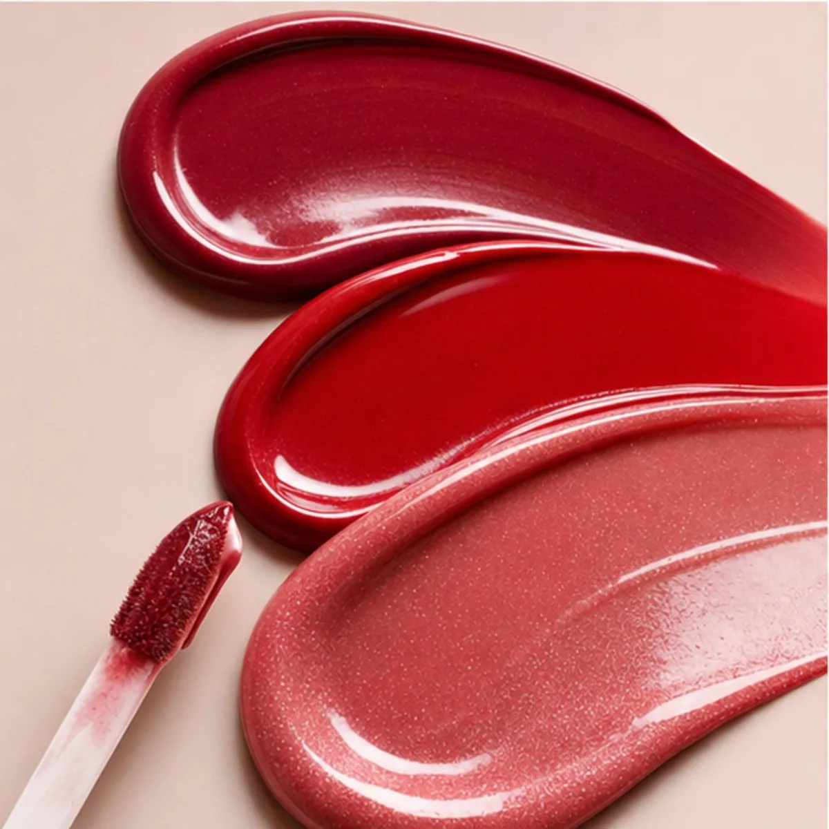 Red and pink lip gloss swatches with an applicator on a pale pink background.