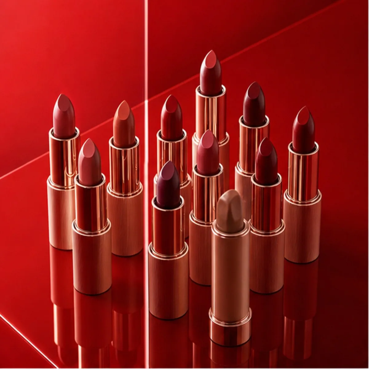Rows of lipsticks in red and nude shades standing on a glossy red surface.